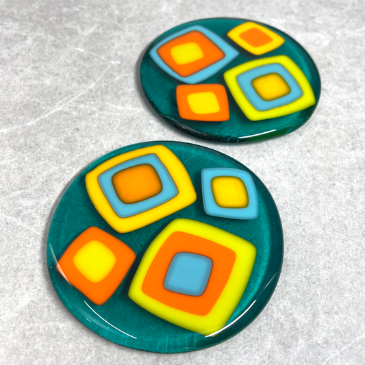 Fused glass round coasters. Size 100mm X 100mm. Transparent turquoise base with orange, lime, turquoise and yellow squares, retro design. Set of 2